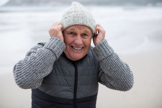 Senior man smiling while standing on the beach during winter. He is wearing a knit hat and grey sweater, enjoying the cold weather by the ocean. Ideal for use in lifestyle, retirement, and outdoor activity themes.