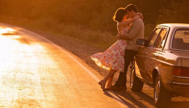 Romantic couple embracing beside vintage sedan on sunlit country road at golden hour