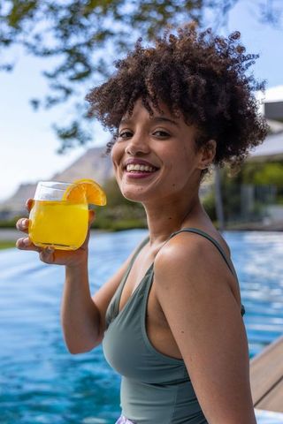 African american woman sipping orange cocktail by poolside on sunny resort deck