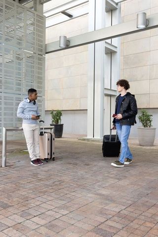 Diverse young travelers standing under modern canopy with rolling suitcases and phones