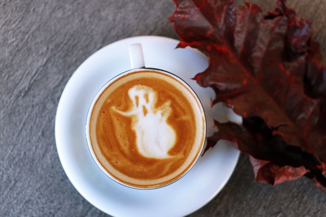 Coffee cup with Ghost art latte on top and autumn leaves on wooden surface. Halloween festivity and autumn season concept