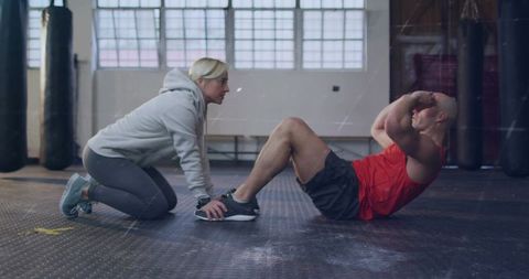 Trainer supporting client doing situps while holding feet in industrial boxing gym
