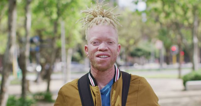 Portrait of a smiling man with albinism, standing in a park filled with greenery. He wears a casual jacket and carries a black laptop bag, indicating he's possibly on his way to work or study. Light filters through the trees, creating a serene and inviting atmosphere. Useful for concepts related to confidence, diversity, young adults, and outdoor activities.
