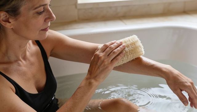 Woman exfoliating forearm with natural loofah while soaking in bathtub sunlight relaxing