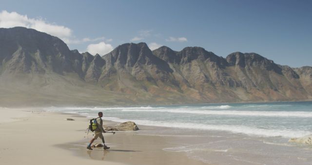 Biracial man with prosthetic leg trekking with backpack on a beach by mountains and the sea. Long distance walking, fitness, challenge, disability, nature and healthy outdoor lifestyle.