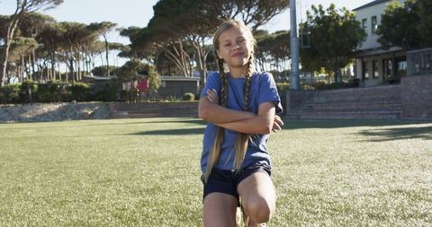 Confident teenage girl kneeling on athletic field in blue shirt and shorts, arms crossed
