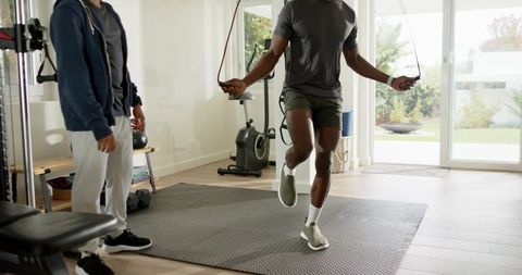 African american man jumping rope for cardio in bright home gym while trainer observing