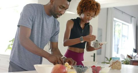 Diverse couple preparing healthy breakfast bowls with banana, oats and berries in kitchen
