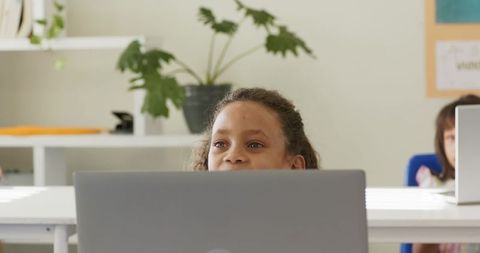 Curious diverse female students peeking over laptops in bright classroom learning moment
