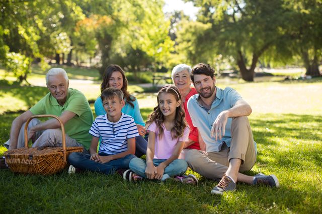 A cheerful multigenerational family is sitting on the grass in a park on a sunny day. They are smiling and enjoying each other's company, with a picnic basket nearby. Perfect for concepts related to family bonding, outdoor activities, leisure time, and healthy lifestyles.