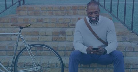 Mature urban cyclist sitting on brick steps checking phone with bicycle and shoulder bag