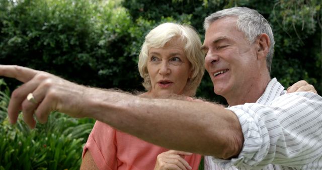 A senior Caucasian couple enjoys a moment outdoors, with the man pointing at something in the distance, with copy space. Their expressions suggest a shared experience or discovery, adding a sense of companionship and curiosity to the image.