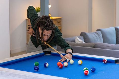 African american young man aiming cue on blue-felt pool table in cozy modern living room