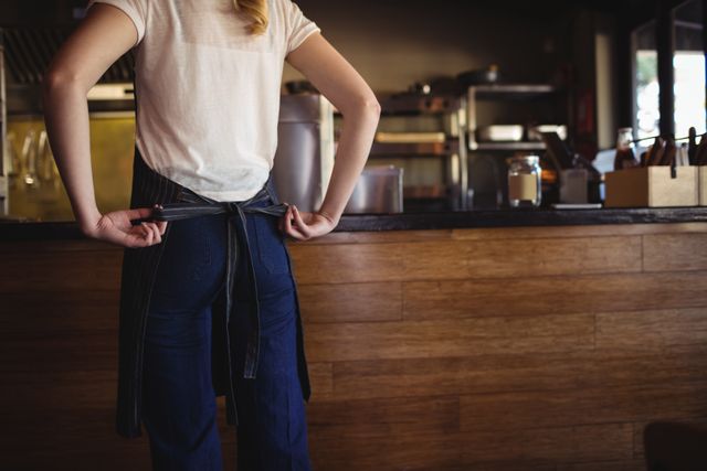 Waitress tying apron in restaurant kitchen, preparing for work. Ideal for illustrating themes of hospitality, food service, restaurant staff, and preparation. Useful for articles, blogs, and advertisements related to the restaurant industry, employee training, and customer service.