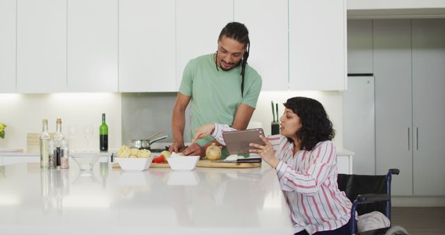 Happy biracial woman in wheelchair using tablet, preparing food with male partner in kitchen. wellbeing and domestic lifestyle with physical disability.