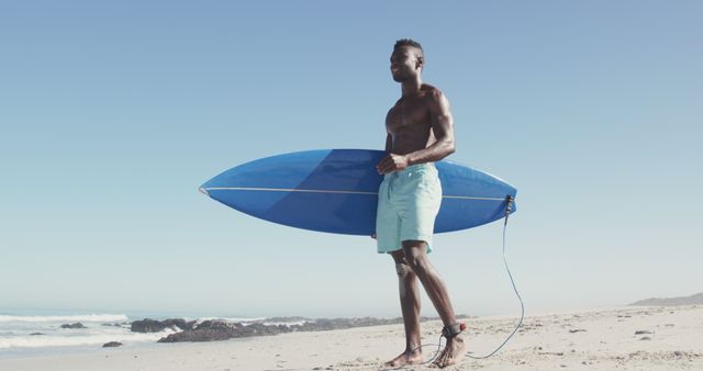 Man holding surfboard standing on sandy beach near ocean waves on a sunny day. Perfect for content related to surfing, coastal lifestyle, outdoor activities, summer vacations, fitness, and seaside adventures.