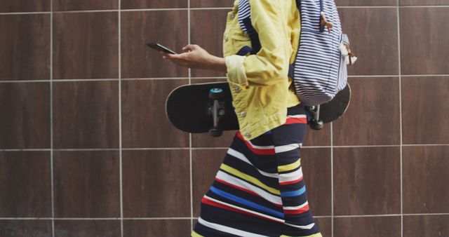 Woman walking in urban environment holding skateboard and smartphone. Dressed in colorful striped skirt, yellow jacket, and striped backpack. Suitable for depicting modern lifestyle, urban fashion, youth culture, and street style themes.