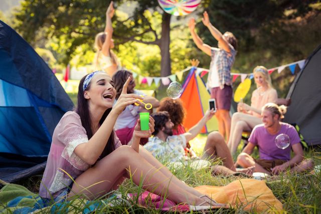 Group of friends enjoying a sunny day at a campsite. One woman is blowing bubbles while others are relaxing and having fun. Ideal for use in advertisements for outdoor activities, camping gear, summer festivals, or travel destinations. Perfect for illustrating themes of friendship, leisure, and outdoor adventures.