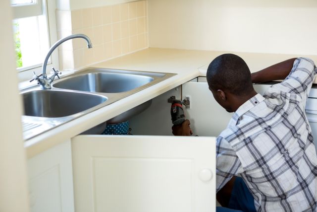 Man repairing kitchen sink at home, ideal for illustrating home maintenance, plumbing services, DIY projects, and handyman work. Useful for blogs, articles, and advertisements related to home repair, plumbing services, and household maintenance.