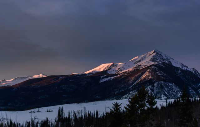 Snow-covered peaks in the Rocky Mountains are illuminated by the warm colors of the setting sun. A tranquil evening sky casts a peaceful light over the snowy landscape, creating a serene and picturesque scene. Ideal for nature enthusiasts, travel promotions, outdoor adventure advertisements, or winter tourism campaigns.