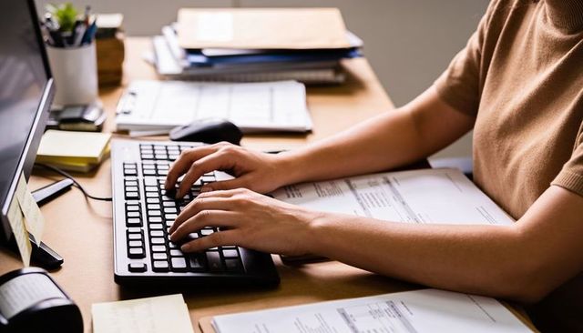 Typing hands on keyboard at organized office desk with printed documents and monitor