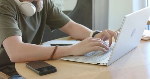 Young man typing on silver laptop at wooden table wearing headphones and smartwatch