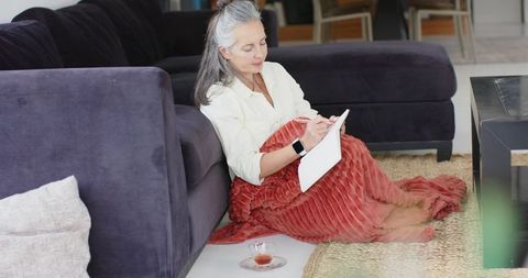 Senior woman journaling on floor wrapped in rust-red blanket leaning on sofa cozy self-care