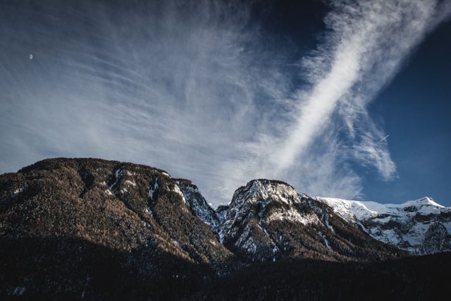 This image depicts majestic snow-capped mountains with dynamic clouds streaking across the sky. Ideal for use in travel brochures, nature documentaries, and outdoor adventure promotions. It beautifully highlights the stark contrast between the snow-covered peaks and the clear sky, capturing the essence of an untouched alpine wilderness.