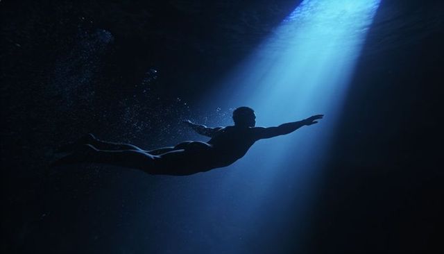 Gliding male silhouette reaching toward column of blue light underwater with rising bubbles