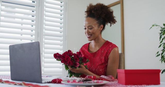 Young woman in red dress arranging red roses on desktop in home office. Perfect for use in content related to Valentine's Day, remote work, floral arrangements, romantic celebrations, and holiday decorations.