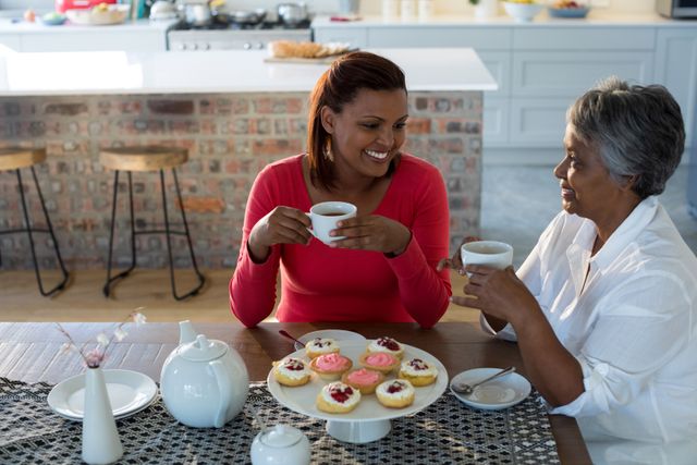 Mother and daughter are sitting at a dining table, enjoying tea and cupcakes in a cozy home kitchen. They are smiling and engaging in a warm conversation, showcasing a strong family bond. This image is perfect for use in advertisements, blogs, and articles about family relationships, home life, and intergenerational connections.