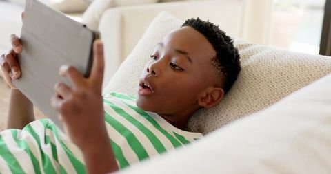 African american boy reclining on sofa using tablet in sunlit modern living room