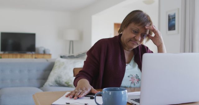 This visual shows an older woman working at home with a laptop, documents, and a coffee cup in her living room. Her expression is stressed as she contemplates her work. This image can be used in articles or advertisements related to remote work challenges, stress management for seniors, and work-life balance.