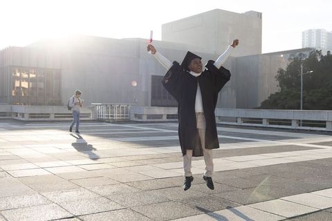 African american graduate jumping on urban campus plaza holding diploma under backlit sun