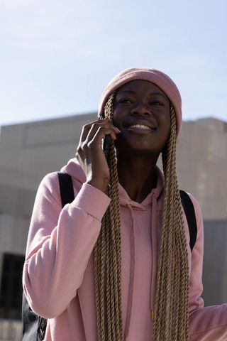 Young woman wearing pink beanie and hoodie talking on phone with backpack in city