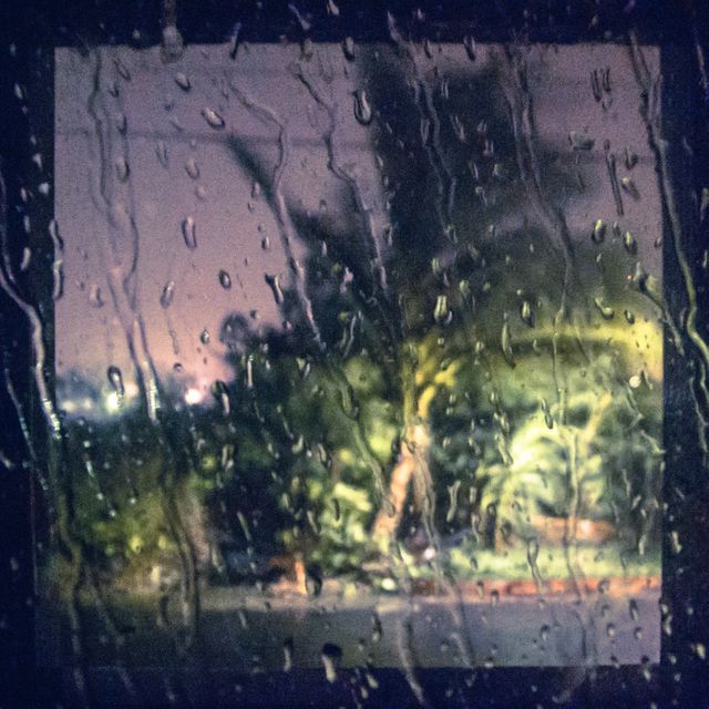 Blurry view through a rain-covered window at night. Raindrops cover glass, giving a distorted view of street with illuminated palm tree. Ideal for use in weather-related themes, background for discussions on storms, ambiance for moody scenes, or illustrating concepts of melancholy and solitude.