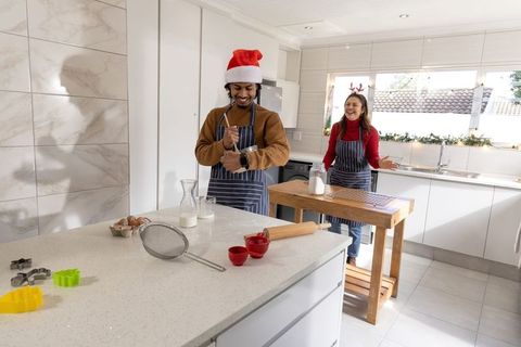 Diverse couple baking christmas cookies in sunlit modern kitchen wearing santa hat, reindeer ears