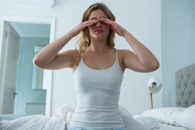 Young woman waking up in bedroom, stretching and covering eyes. Ideal for use in articles or advertisements related to morning routines, sleep health, lifestyle, and home living. Can be used in blogs, social media posts, or wellness websites.