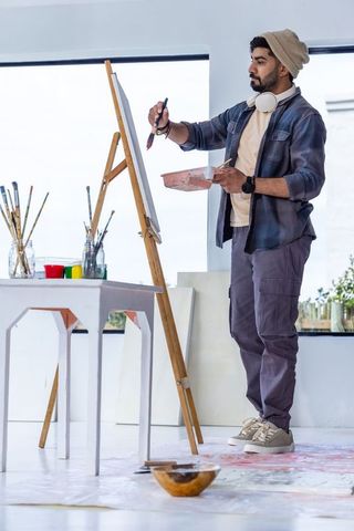 Urban artist painting on wooden easel in sunlit studio wearing beanie and headphones