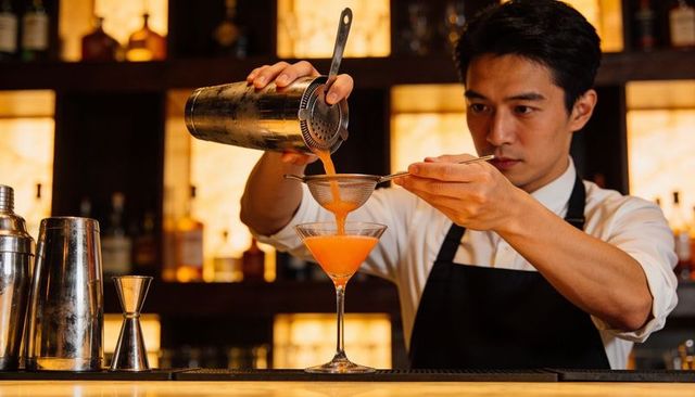 Bartender pouring and straining vibrant orange cocktail into martini glass at upscale bar