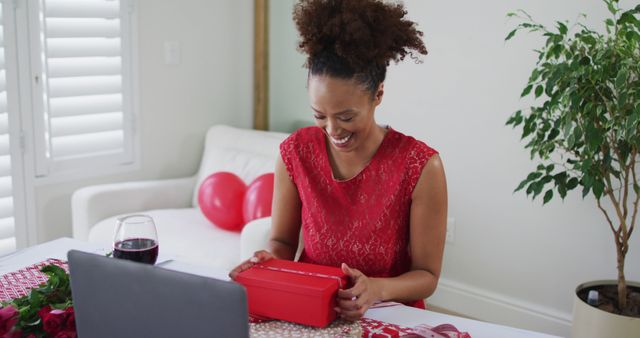 A woman is opening a gift box while smiling, sitting in a bright, modern living room with festive decorations and a glass of wine. This can be used for marketing home celebrations, digital meetups, virtual parties, holiday advertisements, and online gift shop promotions.