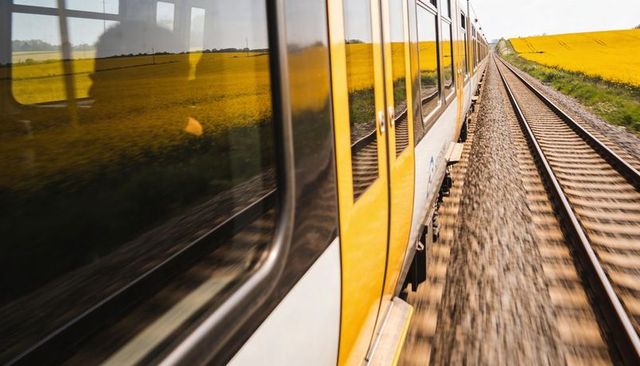 Yellow commuter train speeding past vibrant canola fields along straight railway tracks