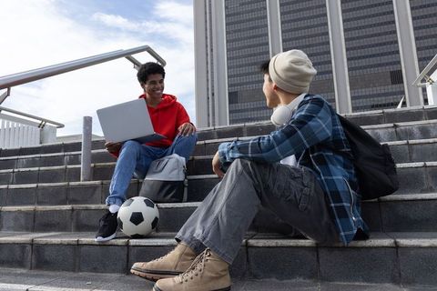 Diverse teenage friends sitting on city steps sharing laptop and soccer ball, chatting