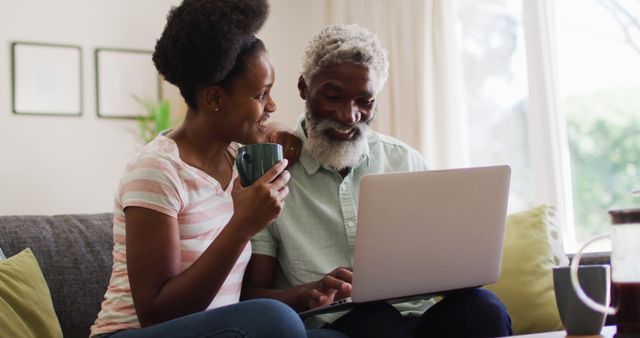 Grandfather and granddaughter are sitting on a couch, smiling and bonding over a laptop while holding coffee mugs. Ideal for content focusing on family relationships, multigenerational activities, and home life. Can be used in advertisements promoting family bonding, technology connecting generations, or home comfort products.
