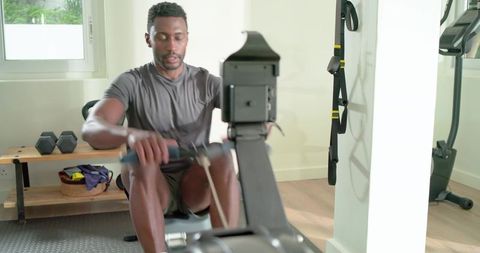 Mid adult african american man rowing on indoor machine in bright minimalist home gym
