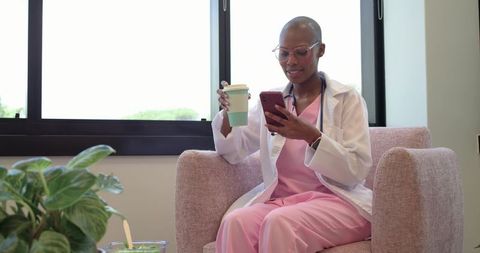 African american nurse checking smartphone during coffee break in bright clinic lounge