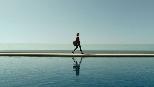 Solo woman walking along glass-railed promenade by infinity pool with reflection