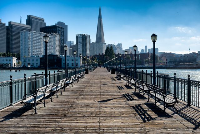 Peaceful view from wooden pier with benches lining path, leading toward skyline with tall buildings. Ideal for websites focusing on urban landscapes, travel destinations or architectural photography. Use in blogs, advertisements, or social media to convey serenity, city life, and urban beauty.
