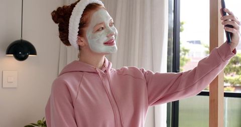 Young woman taking selfie with green facial mask and headband in cozy pink hoodie