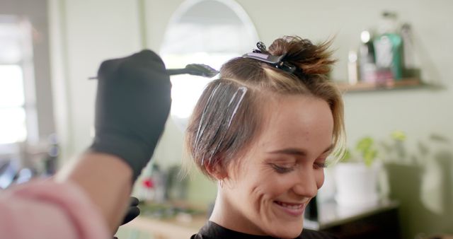 Young woman looking pleased as she gets her hair colored at a salon. Ideal for use in beauty, fashion, lifestyle, cosmetic, and professional services marketing. Displays professional hairdressing techniques and customer satisfaction.